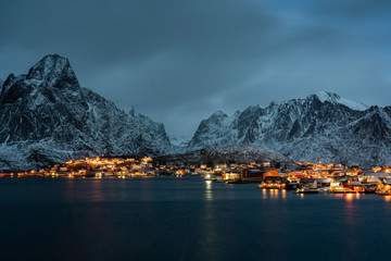Reine village harbor at night with building's lights and outstanding snowy mountains. Lofoten islands, Norway.