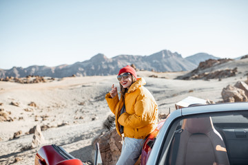 Young woman dressed in bright jacket and hat enjoying road trip on the desert valley, standing near...