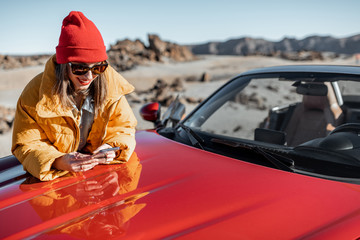 Young woman dressed in bright jacket and hat enjoying road trip on the desert valley, standing with...