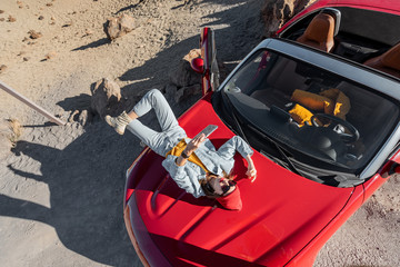 Lifestyle portrait of a young woman enjoying road trip on the desert valley, lying on the car hood and photographing on phone. View from above