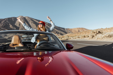 Carefree woman dressed casually in jeans and red hat enjoying road trip on the volcanic valley, raising hands from convertible car on the roadside