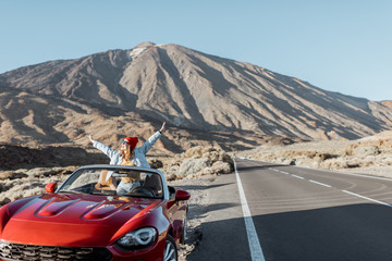 Carefree woman enjoying road trip on the volcanic valley, raising hands from convertible car on the...