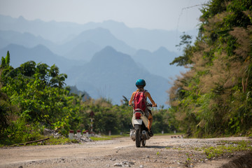 Young woman rides the scooter on an empty road in tropics with mountains on the background