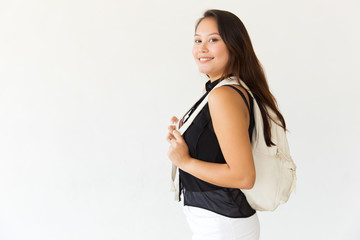 Female student with backpack smiling at camera. Side view of cheerful young Asian woman with backpack standing and looking at camera on white background. Education concept