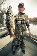 Naklejka premium Young amateur angler holds the big Carp fish (Cyprinus carpio) being in the boat on the river