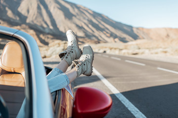 Young woman traveling by convertible car on the desert valley, pulling legs out the car window on...