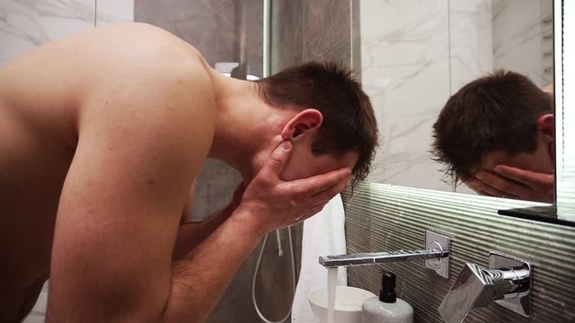 Topless handsome man washing face in sink under running cold water from faucet. Muscular man doing morning routine clean procedures. Looking to the mirror reflection and smiling. Slow motion