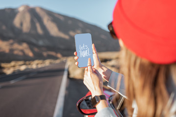 Woman traveling by car on the volcanic valley, leaning out of the car window, holding smartphone...