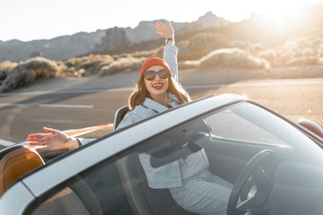 Lifestyle portrait of a playful woman having fun during a road trip, raising hands out of the...