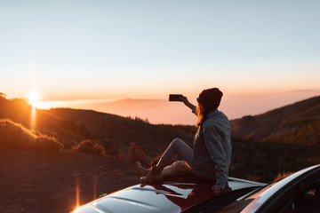 Young woman photographing with phone beautiful landscape during a sunset, sitting on the car hood while traveling high in the mountains