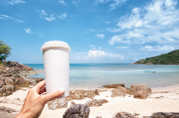 Hand holds a white cardboard paper mug for takeaway drinks, against  beautiful sea shore background, environment concept, mock up