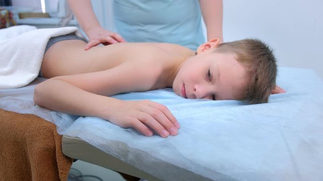 Doctor massagist making therapeutic massage to teen boy on spine back in clinic. Child boy lying on couch and talking. Cure massage, rehabilitation after injury. Medicine treatment for children.