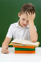 The boy is engaged in school, sitting on the background of a green blackboard and reading a book