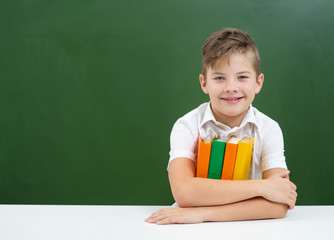 The boy is engaged in school, sitting on the background of a green blackboard with a stack of books in an embrace