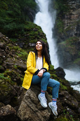 Naklejka na ściany i meble The girl tourist in a yellow jacket enjoys freedom against the backdrop of the waterfall. Young woman looking on the majestic waterfall in the Norway. Travelling, lifestyle, adventure, concept.