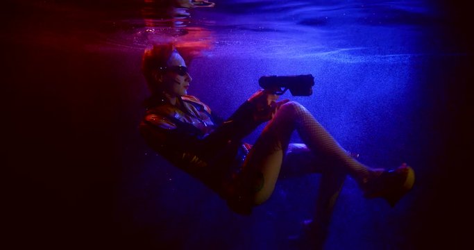 young woman with gun blaster in hands is floating underwater in dark aquarium, subaquatic shot