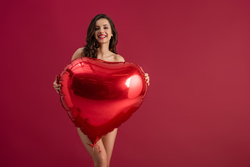 sexy girl smiling at camera while holding heart-shaped balloon isolated on red