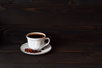 a white Cup with poured black coffee and saucer stands on a dark wooden background. coffee beans are scattered on a saucer along with star anise.