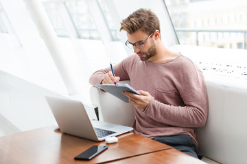 Image of man using laptop and clipboard while working in cafe indoors