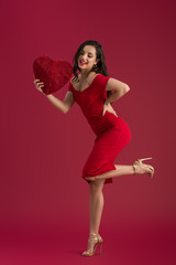 smiling, elegant girl holding decorative heart while standing on one leg on red background