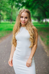 Portrait of a charming blond woman wearing beautiful white dress standing on the road under trees.