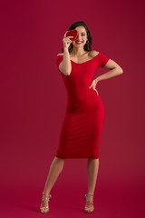 sensual, elegant girl covering eyes with heart-shaped gift box while standing with hand on hip on red background