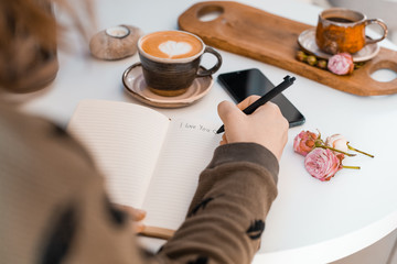 Girl writes I love you in a notebook on a table next to a phone and a cup of coffee decorated with a heart and rosebuds
