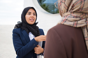 Confident Muslim businesswoman talking with coworker on street. Focused young woman holding documents and gesturing. Business and communication concept