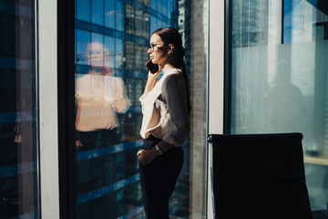 Confident businesswoman talking on smartphone beside office window