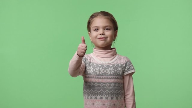 Little happy actress 5 years old with brown hair dressed in pink sweater shows thumb up. Sweet smiling girl in earrings walks around the studio. Stays on green background, chroma key.