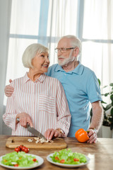 Selective focus of senior man hugging smiling wife while cooking on kitchen table