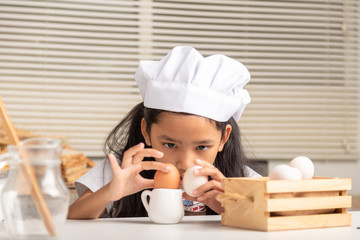 A little Asian girl wearing a white chef hat is staring at eggs place on the small jug