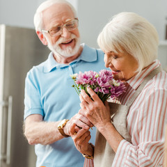 Selective focus of senior woman in apron smelling bouquet by smiling husband in kitchen