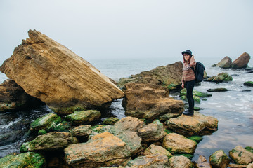 Obraz premium Young woman tourist in hat and with backpack standing on a rock against a beautiful sea, looking at sea, on horizon. Place for text or advertising