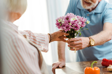 Cropped view of senior man giving bouquet to wife beside fresh vegetables on kitchen table