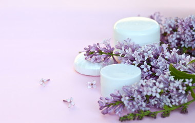 Obraz na płótnie Canvas Two jars of moisturizer for face and body care and a cosmetic soap with sprigs of blooming lilacs on a pink background. Spring romantic still life. Close-up, blanc, mock up, macro, place for text