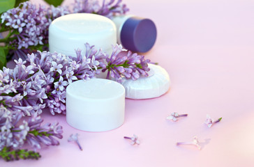 Obraz na płótnie Canvas Cosmetic moisturizers creams for face and body and a fragrant soap with sprigs of blooming lilacs on a pink background. Spring tender still life. Close-up, shallow depth of field, place for text