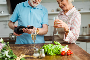 Selective focus of smiling woman looking at husband pouring champagne by bouquet and vegetables on kitchen table