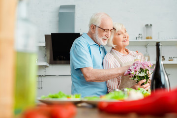 Selective focus of smiling senior couple with bouquet and champagne hugging beside vegetables on kitchen table