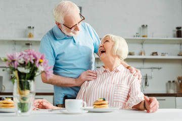 Selective focus of senior woman laughing while looking at husband by coffee and pancakes on table...