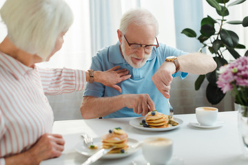 Fototapeta na wymiar Selective focus of senior woman touching husband with pancakes by coffee and flowers on table