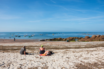 Fototapeta na wymiar Normandy, France. Senior people (back view) relax at sandy beach, sunbath and admire beautiful bay with mooring fishing boats. Summer seaside vacation background. Lifestyle, retired wellness concepts