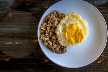 Fried Pork with Garlic, Pepper and fried egg on rice on white dish and on wooden table. Image on top view.