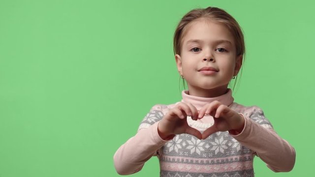 Video portrait of happy baby girl 6 years old in studio shows heart gesture by two hands, looking at camera. Sweet caucasian baby dressed in pink sweater stays on green chroma key background.