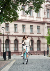 Beautiful young girl in white dress enjoying her summer day riding a bike, concrete beauty, purple building on the background, healthy lifestyle in town