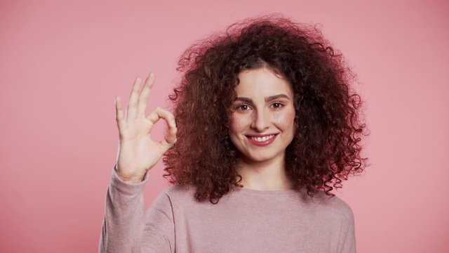 Winner. Success. Positive girl making OK sign over pink background and smiles to camera. Body language. Young curly woman with trendy glitter freckles make-up