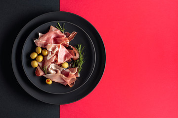 Prosciutto with rosemary and green olives on a black plate. Top view.