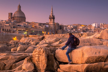 Man sitting on the rock and watch beautiful architecture of the Valletta city at dawn, Malta - obrazy, fototapety, plakaty