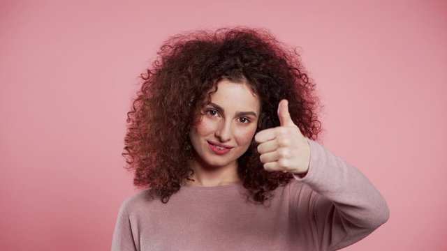 Winner. Success. Positive girl making thumbs up sign over pink background and smiles to camera. Body language. Young curly woman with trendy glitter freckles make-up