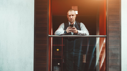 A handsome mature elegant hispanic businessman is pensively chatting using his smartphone while standing indoors on the balcony of a modern office; a copy space area on the left for a message or logo